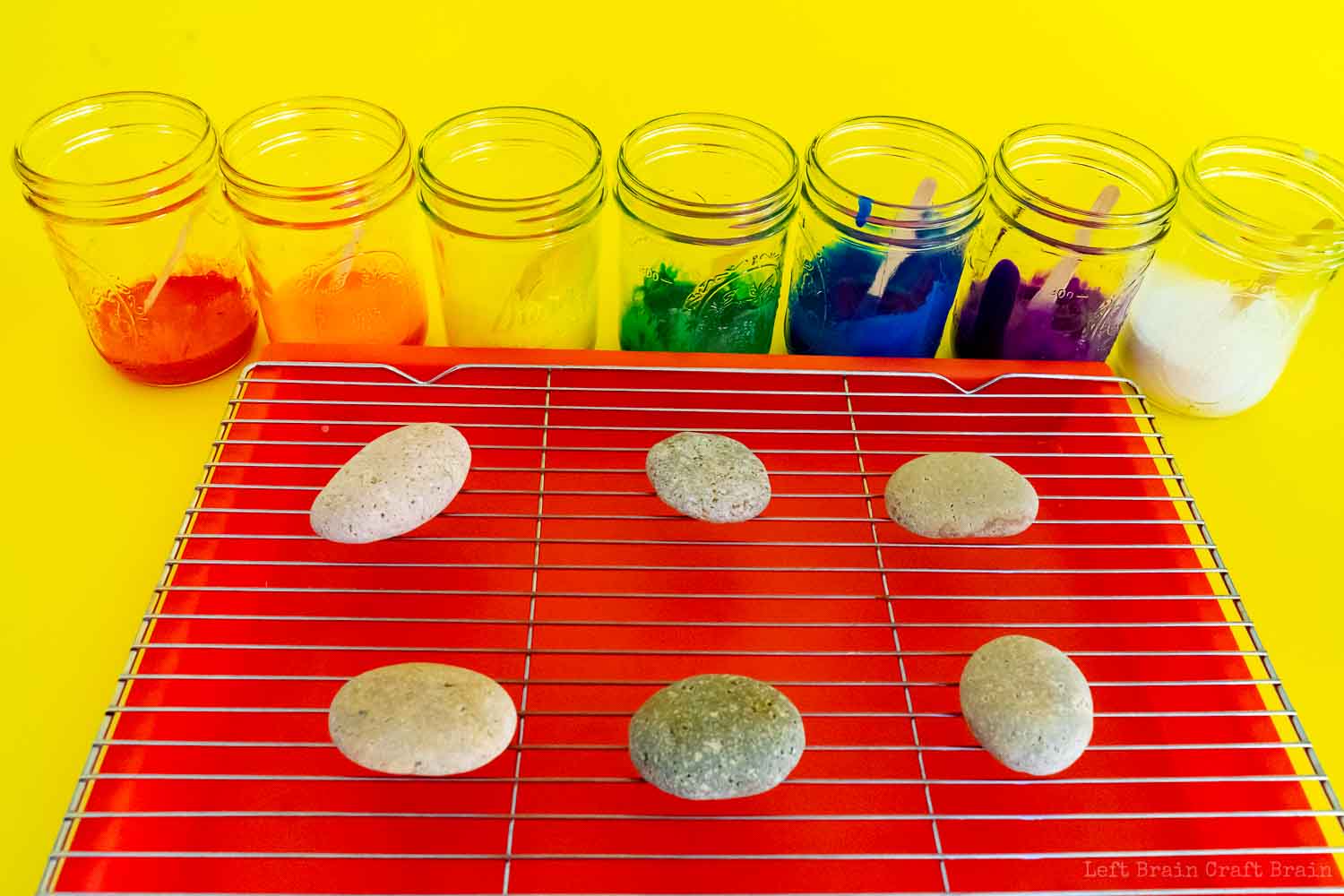 rocks on wire rack on tray in front of jars with different colors of paint in rainbow